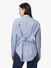 Load image into Gallery viewer, A woman shown from the back wearing a light blue and white vertically striped button-up shirt with a tie detail at the waist, paired with dark blue jeans, against a plain light gray background.
