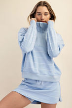 Load image into Gallery viewer, A woman modeling a light blue matching lounge set consisting of a half-zip sweatshirt and a drawstring mini skirt, both with thin white horizontal stripes, against a solid beige background.
