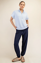 Load image into Gallery viewer, A woman modeling a light blue short-sleeve polo shirt and navy jogger pants with brown double-strap sandals, standing against a plain beige studio background.
