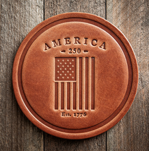 Load image into Gallery viewer, Brown leather coaster with 'America 250' and American flag design on a wooden background

