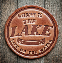 Load image into Gallery viewer, Round brown leather coaster with 'Welcome to the Lake Life on the Water' text on a wooden background
